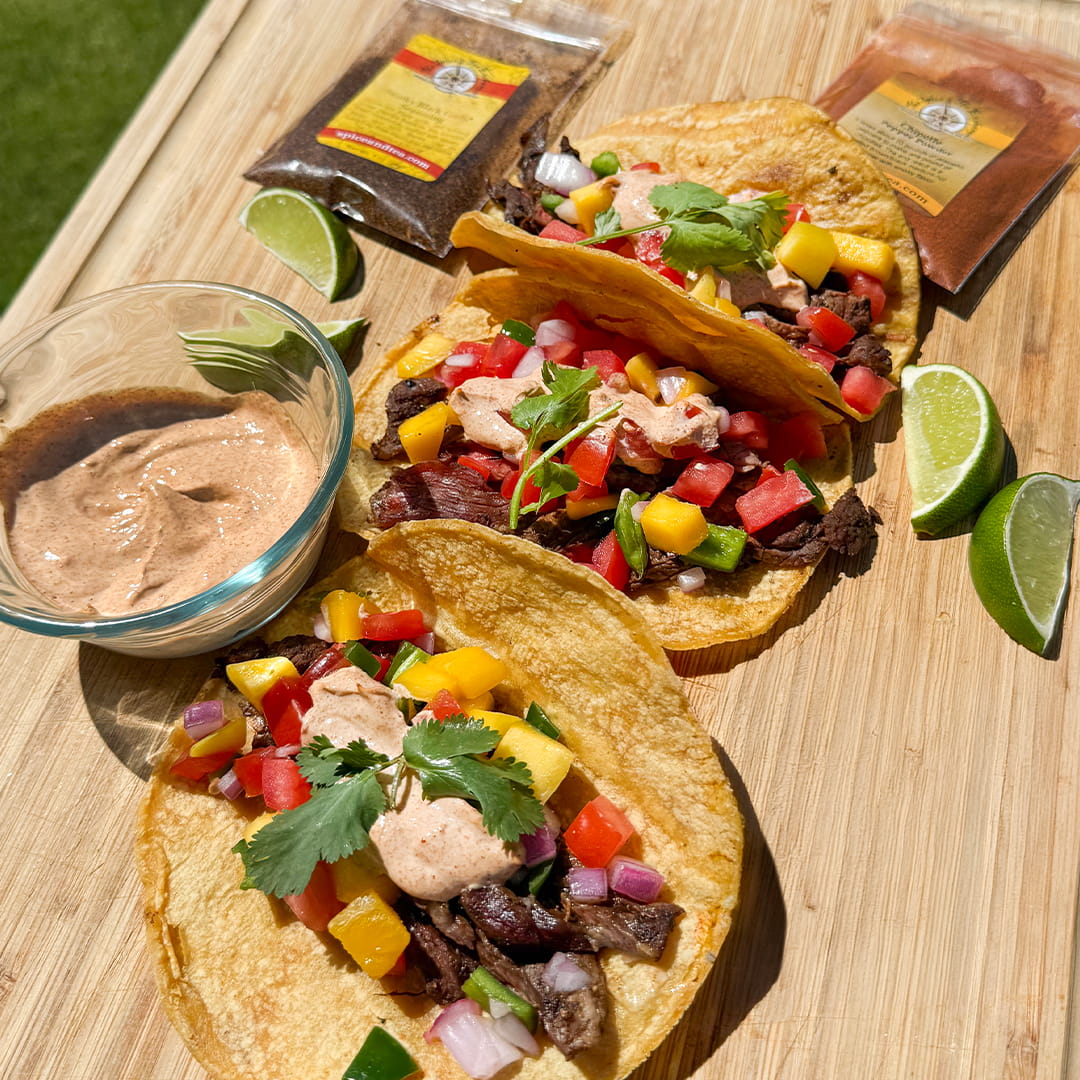 Three Steak Tacos (Kit), featuring grilled meat, diced veggies, and chipotle honey crema, are arranged on a wooden board alongside lime wedges, a bowl of sauce, and two seasoning kit spice packets. Bright daylight emphasizes the fresh ingredients.